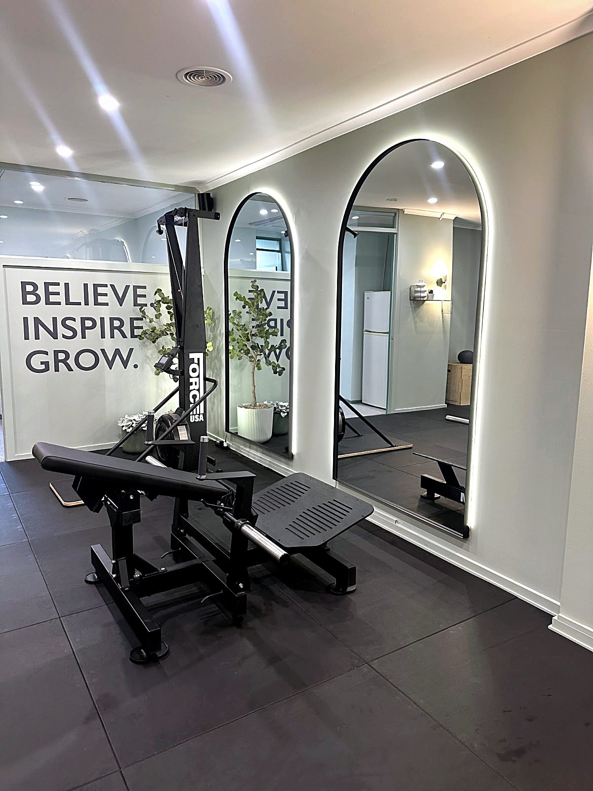 Backlit arched mirrors and the chest press beside the Believe Inspire Grow wall