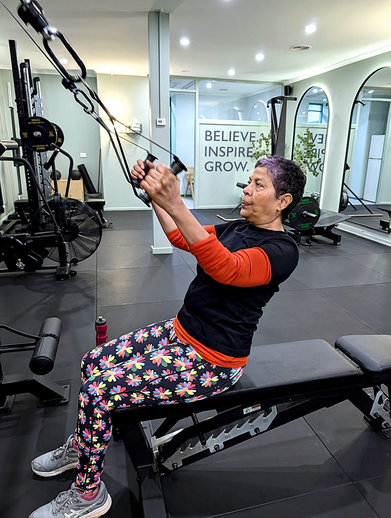 A client in her sixties pressing on the cable machine, with the Believe Inspire Grow wall behind her