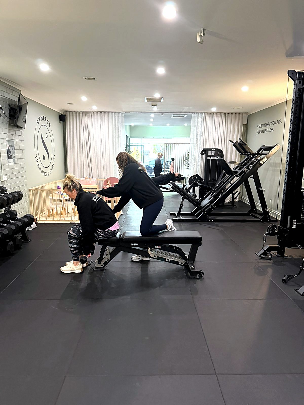 Two Synergy coaches guiding clients through their programs on the studio floor, beside the playpen